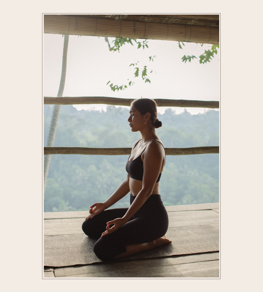 Woman meditating in black activewear on a wooden deck overlooking lush jungle.