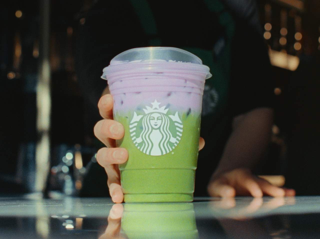 A barista places an Iced Lavender Matcha in a clear Starbucks to-go cup on a reflective counter.